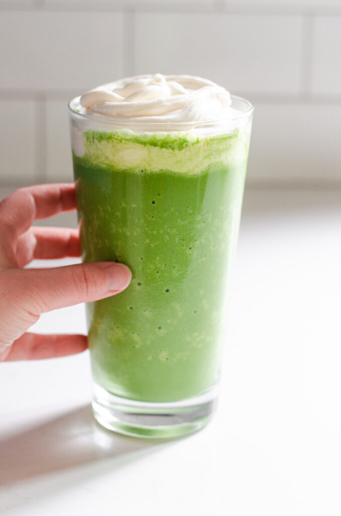 A clear glass filled with a matcha Frappuccino with a hand holding it on the left.