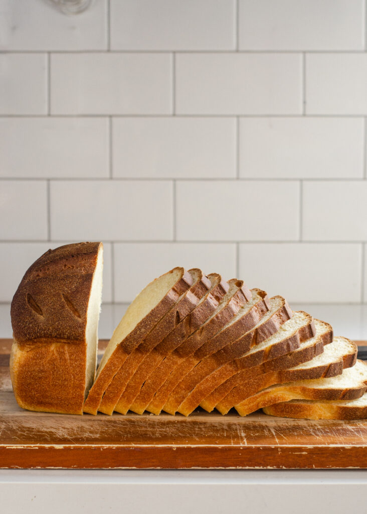 A loaf of sourdough sandwich bread mostly sliced up on a wooden cutting board with a white subway tile backsplash behind it.