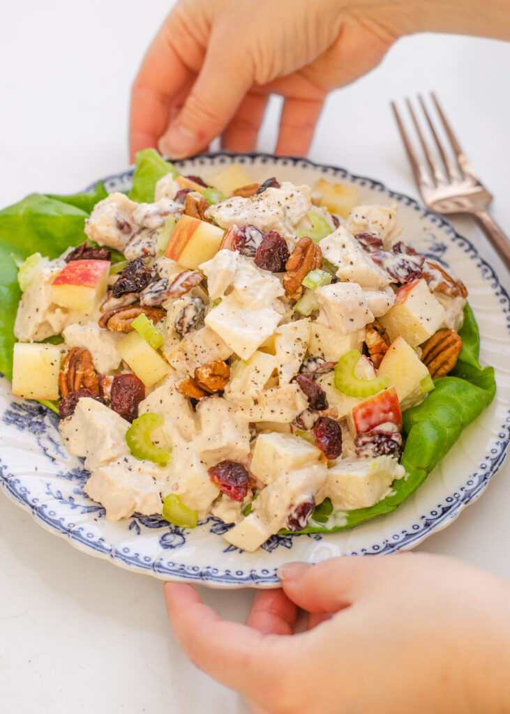 Cranberry pecan chicken salad on top of leaves of butter lettuce on a vintage blue plate with hands on the sides of the plate and a fork in the background.