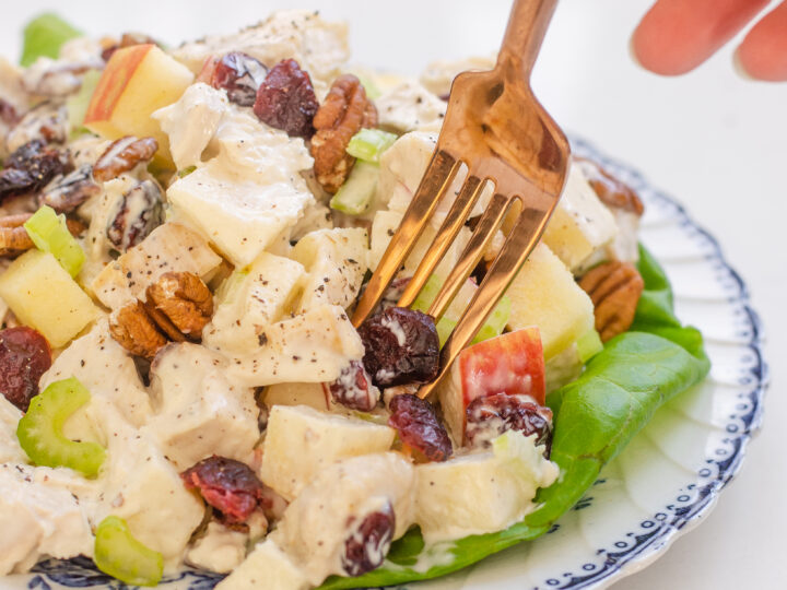 A plate of cranberry pecan chicken salad with lettuce leaves underneath, and a hand poking a fork into the salad.