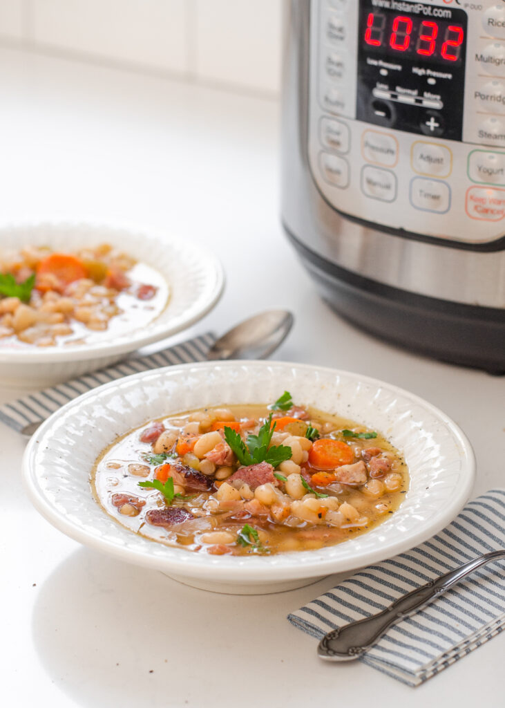 Two bowls of ham and bean soup on a white counter with napkins and spoons to the side with the Instant Pot in the background.