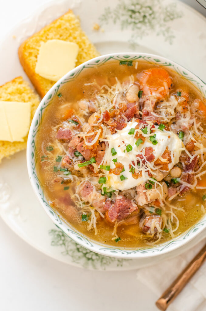 Close up shot of the ham and bean soup with added sour cream, bacon, cheese, and green onions with cornbread off to the side.