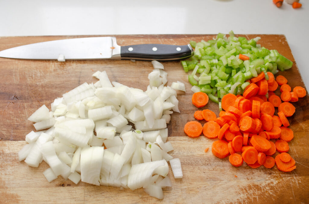 Carrots, celery, and onion on a wooden cutting board all chopped up with a chef's knife in the background.