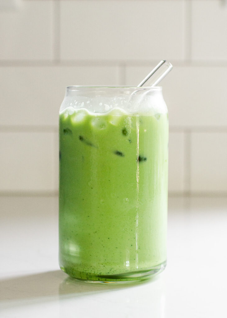 A glass of matcha iced latte on a white countertop with a glass straw in it.