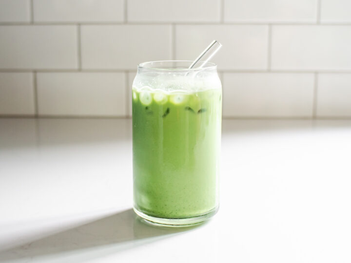 A glass of iced matcha latte on a white countertop with a glass straw in it.