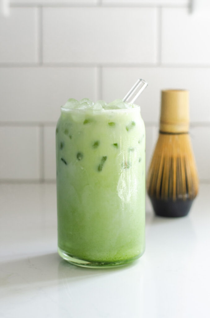 A glass of iced matcha latte on a white countertop with a glass straw in it and a bamboo matcha whisk in the background.