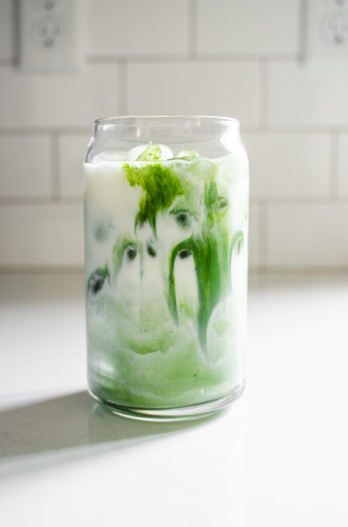 A glass of iced matcha latte on a white countertop with a glass straw in it.