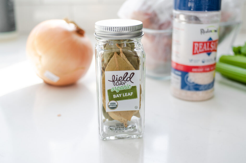 A jar of bay leaves on a white countertop with other ingredients for making chicken bone broth in the background.