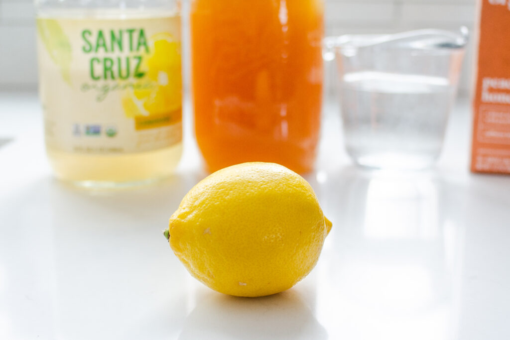 A lemon on a white countertop with lemonade, honey, and water in the background.