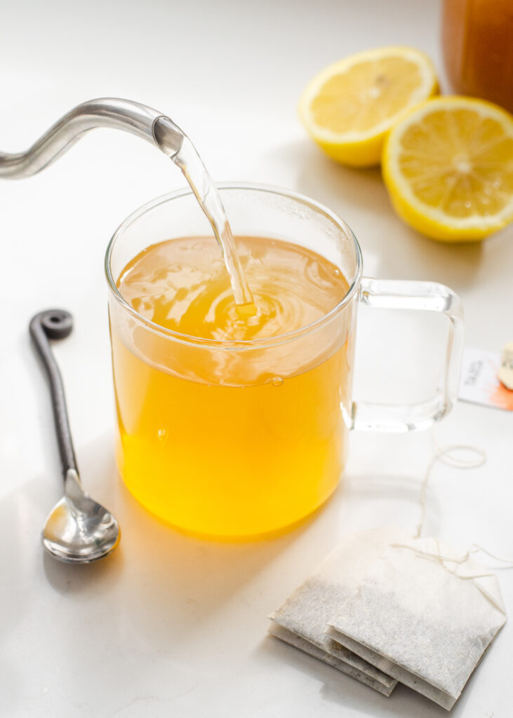 A mug of Starbucks medicine ball tea with a spoon on one side, tea bags on the other, and a lemon behind it, and a tea kettle pouring more water into the mug.
