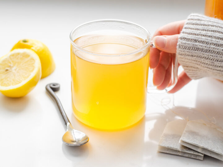 A handing holding the handle of a mug of Starbucks medicine ball tea with a lemon and spoon on one side and tea bags on the other.