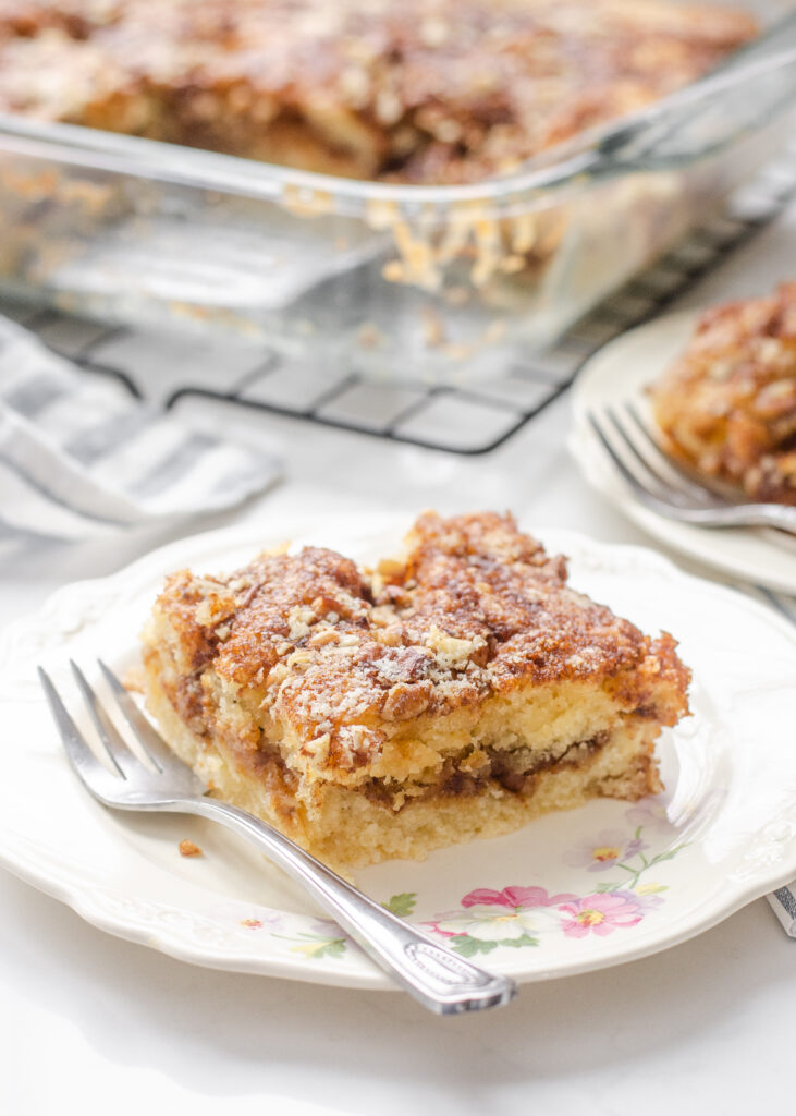 A slice of sourdough coffee cake on a small plate with a fork to the side with the pan in the background on a wire cooling rack.