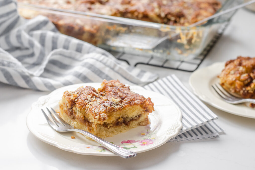 A slice of sourdough coffee cake on a small plate with a fork to the side with the pan in the background on a wire cooling rack.