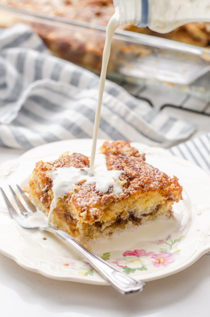 Pouring cream over a slice of sourdough coffee cake on a small plate with a fork to the side with the pan in the background on a wire cooling rack.