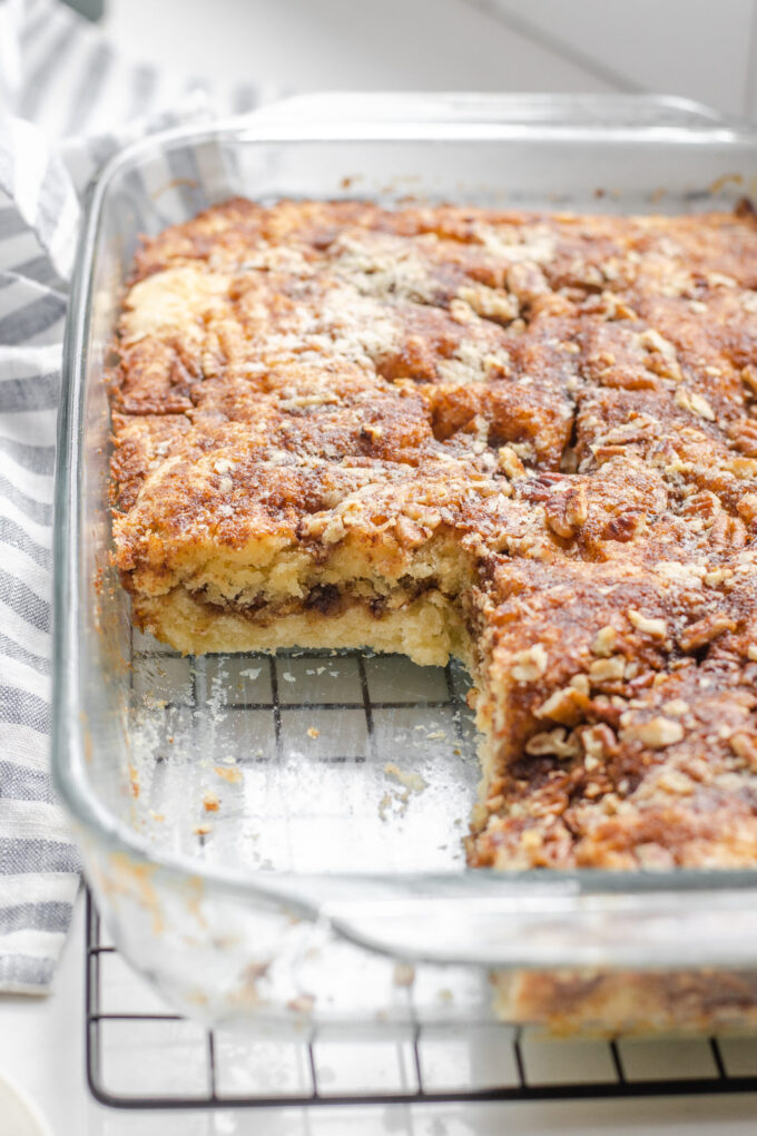 A pan of sourdough coffee cake made with cinnamon and pecans on a wire cooling rack with a linen tea towel in the background, and a piece taken out.