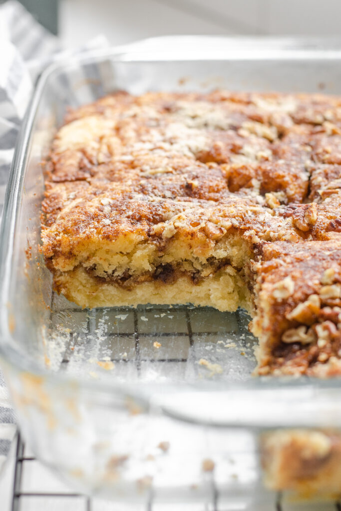 A pan of sourdough coffee cake made with cinnamon and pecans on a wire cooling rack with a linen tea towel in the background, and a piece taken out.