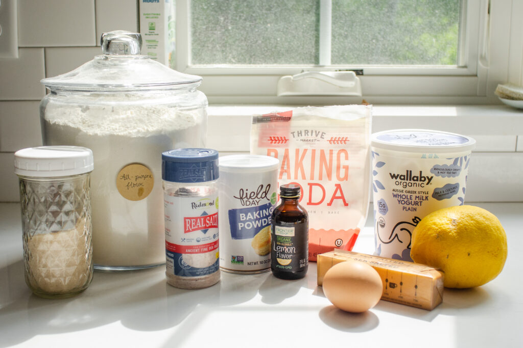 All of the ingredients needed to make lemon scones at home laid out on a white countertop with the sun coming through a window in the background.