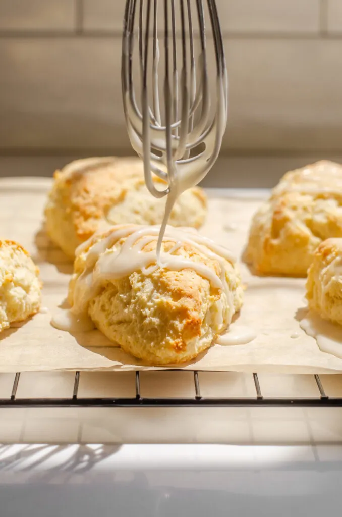 Drizzling a glaze on top of lemon scones on parchment paper on top of a wire cooling rack.