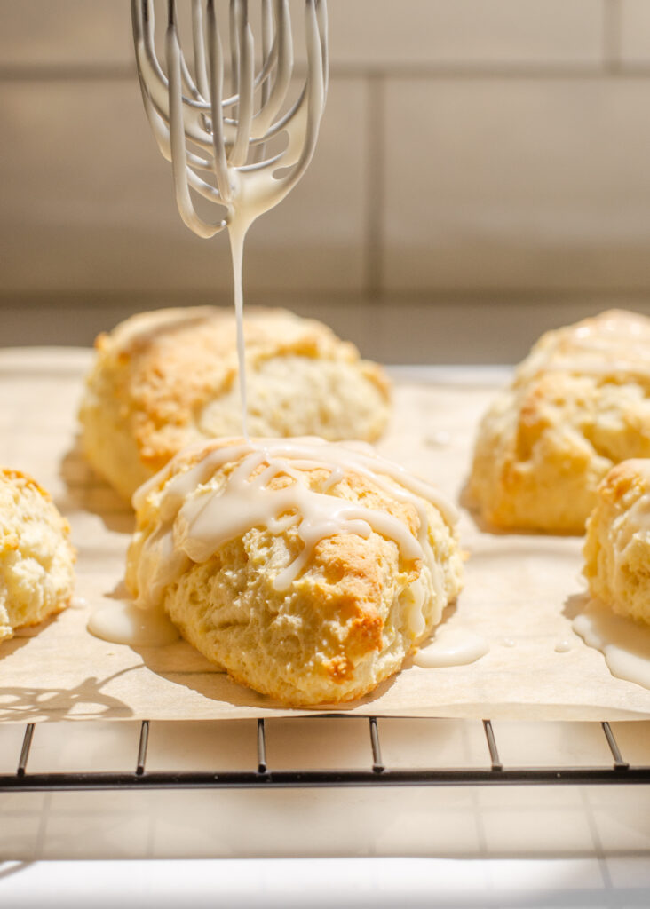 Drizzling a glaze on top of lemon scones on parchment paper on top of a wire cooling rack.