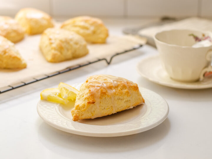 A lemon scone on a plate with small slices of lemon next to it and more scones on a wire rack behind it with a teacup beside it.