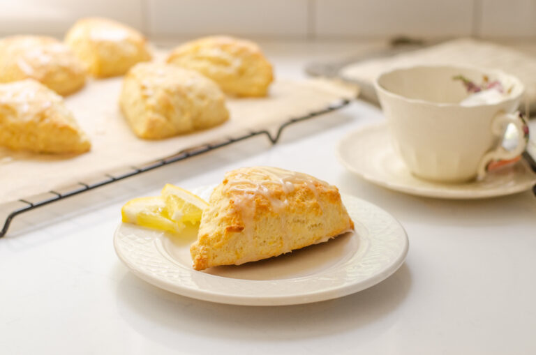 A lemon scone on a plate with small slices of lemon next to it and more scones on a wire rack behind it with a teacup beside it.