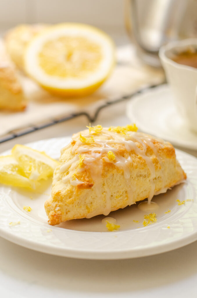 A lemon scone on a plate with small slices of lemon next to it and more scones on a wire rack behind it with a teacup beside it.