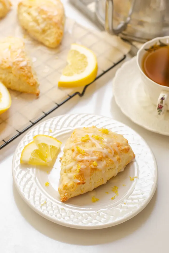 A lemon scone on a plate with small slices of lemon next to it and more scones on a wire rack behind it with a teacup beside it.