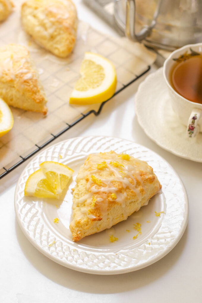 A lemon scone on a plate with small slices of lemon next to it and more scones on a wire rack behind it with a teacup beside it.