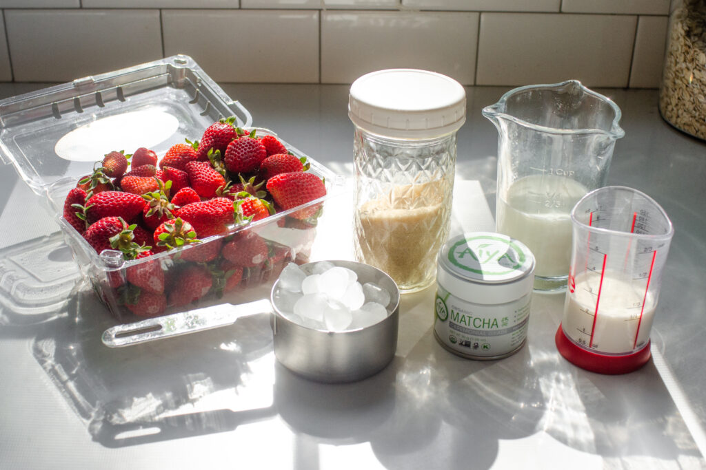 All of the ingredients needed to make an iced strawberry matcha latte laid out on a white countertop with the sun shining through the window.