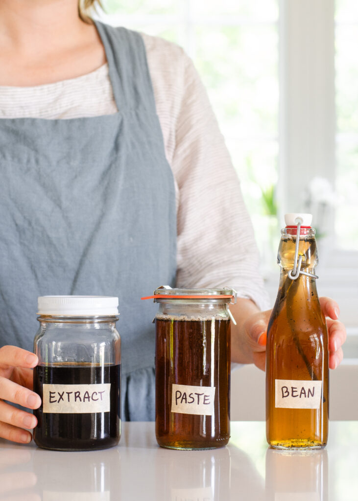 Three bottles of vanilla extract on a countertop with a person in an apron behind them holding two of the bottles.