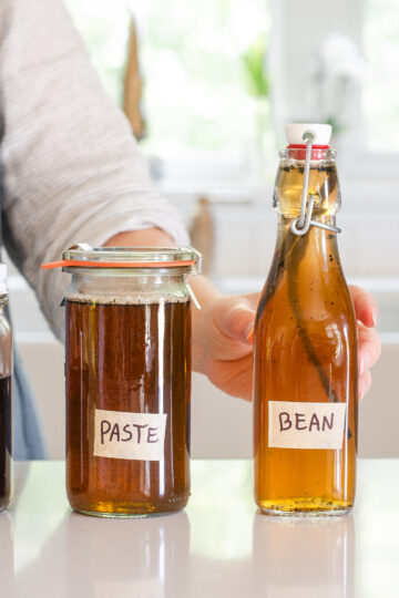 A woman in a linen apron standing behind jars of homemade vanilla syrup for flavoring coffee.