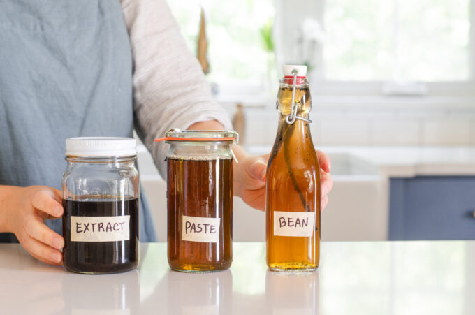 A woman in a linen apron standing behind jars of homemade vanilla syrup for flavoring coffee.