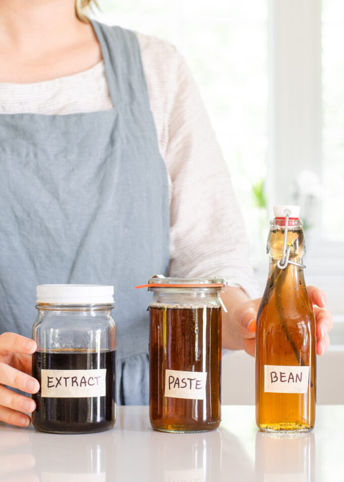 A woman in a linen apron standing behind jars of homemade vanilla syrup for flavoring coffee.