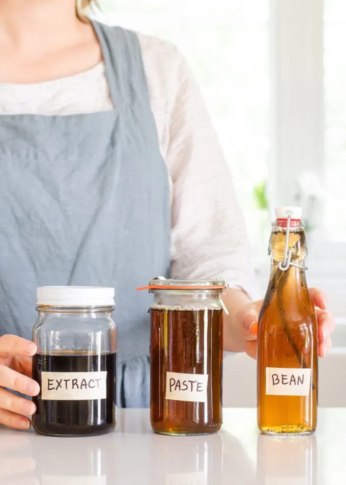A woman in a linen apron standing behind jars of homemade vanilla syrup for flavoring coffee.
