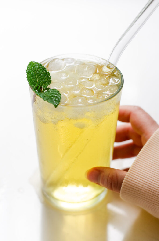 A 12 ounce glass of homemade Starbucks iced green tea with a clear glass straw and a mint leaf with a hand holding the glass.