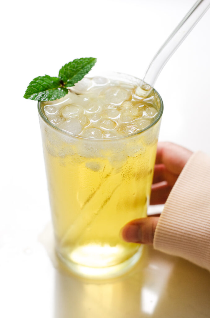 A 12 ounce glass of homemade Starbucks iced green tea with a clear glass straw and a mint leaf with a hand holding the glass.