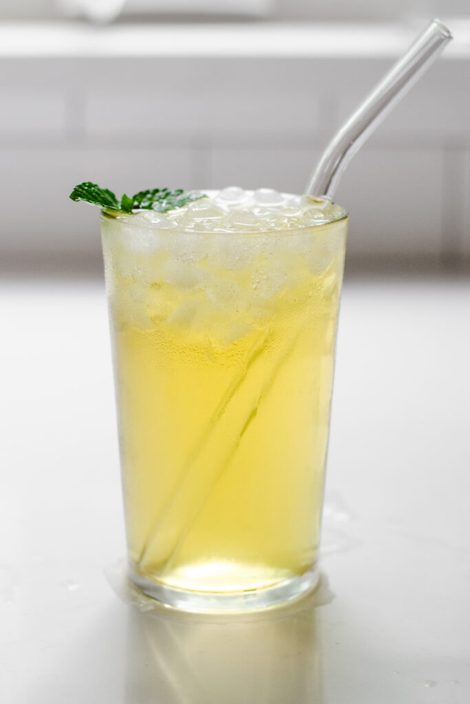 A glass of homemade Starbucks iced green tea with a glass straw sticking out of it and a mint leaf on top.