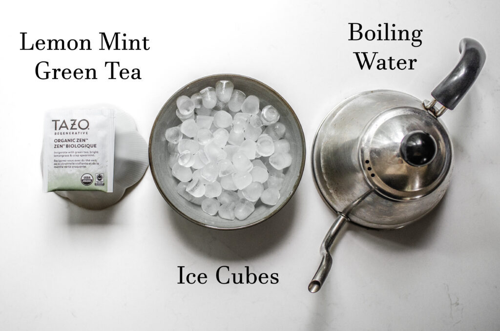 A green tea bag, a bowl of ice, and a tea pot laid out on a white countertop.