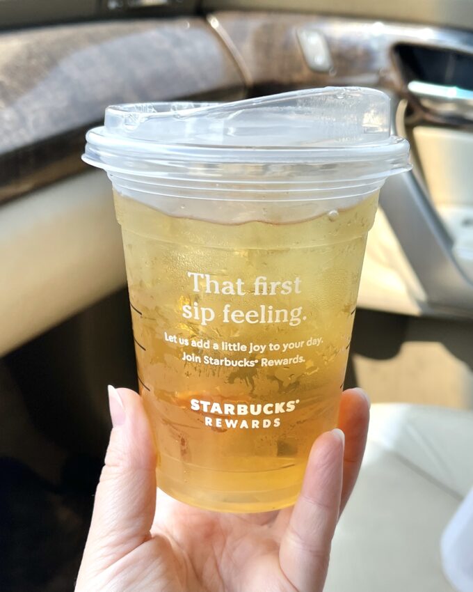 A hand holding a Starbucks cup of iced green tea while sitting in a car.