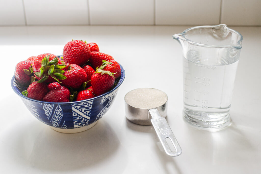 All of the ingredients needed to make strawberry syrup lined up on a white countertop.