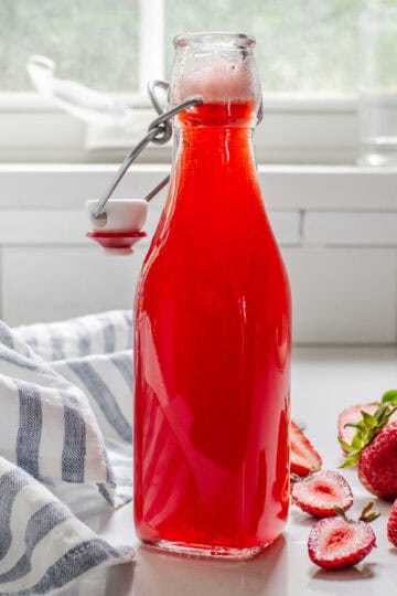 A bottle of strawberry syrup for drinks on a white countertop with fresh strawberries on one side and a blue striped linen napkin on the other side.
