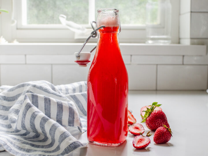 A bottle of strawberry syrup for drinks on a white countertop with fresh strawberries on one side and a blue striped linen napkin on the other side.