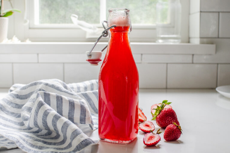 A bottle of strawberry syrup for drinks on a white countertop with fresh strawberries on one side and a blue striped linen napkin on the other side.