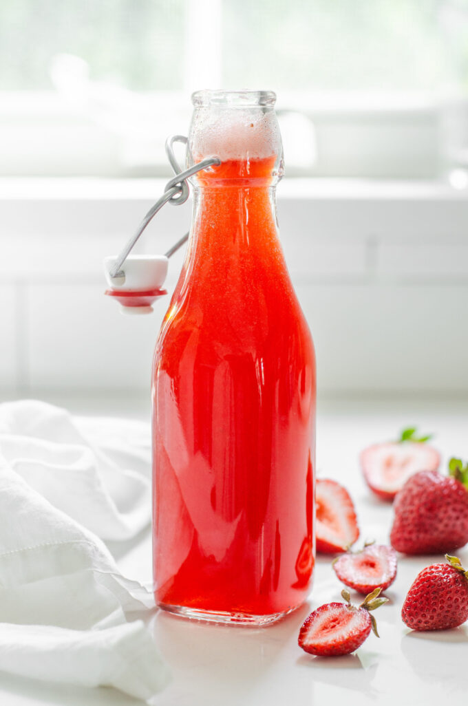 A bottle of strawberry syrup for drinks on a white countertop with fresh strawberries on one side and a blue striped linen napkin on the other side.