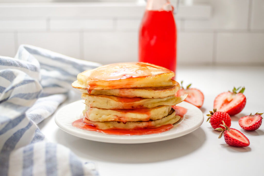 A stack of pancakes on a small plate with strawberry syrup on top.