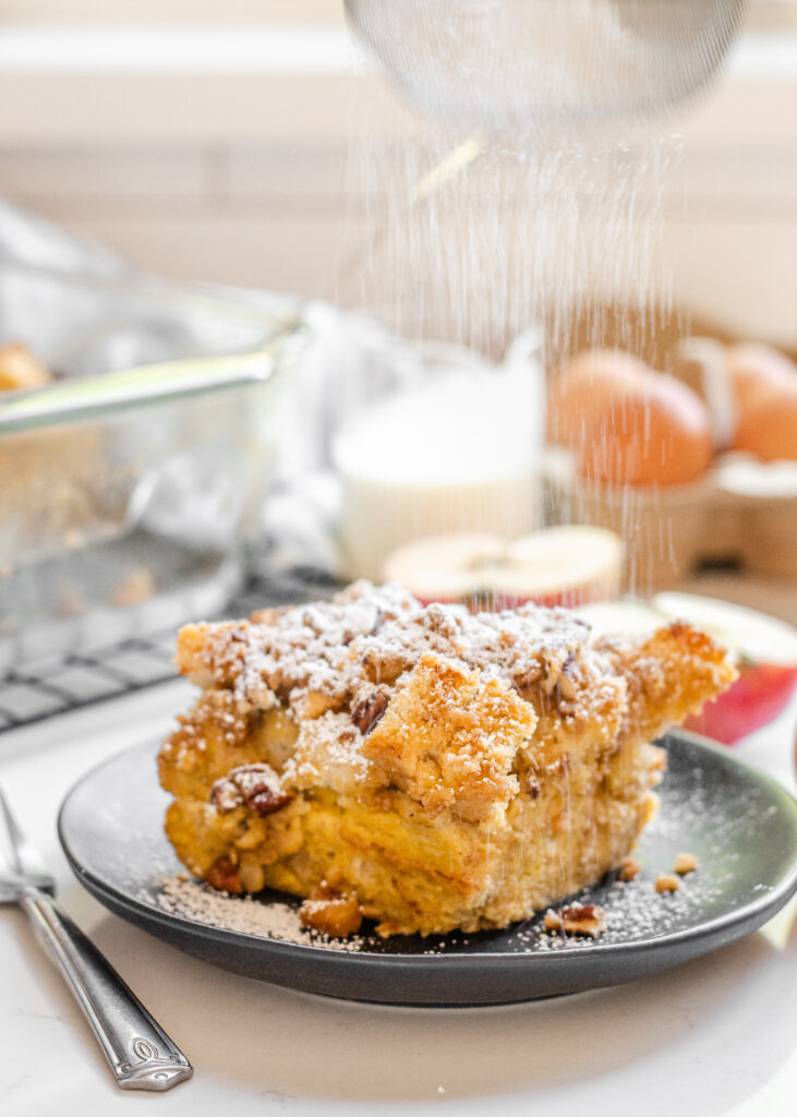 A slice of apple French toast casserole on a grey plate with apples in the background and powdered sugar being sprinkled on top.