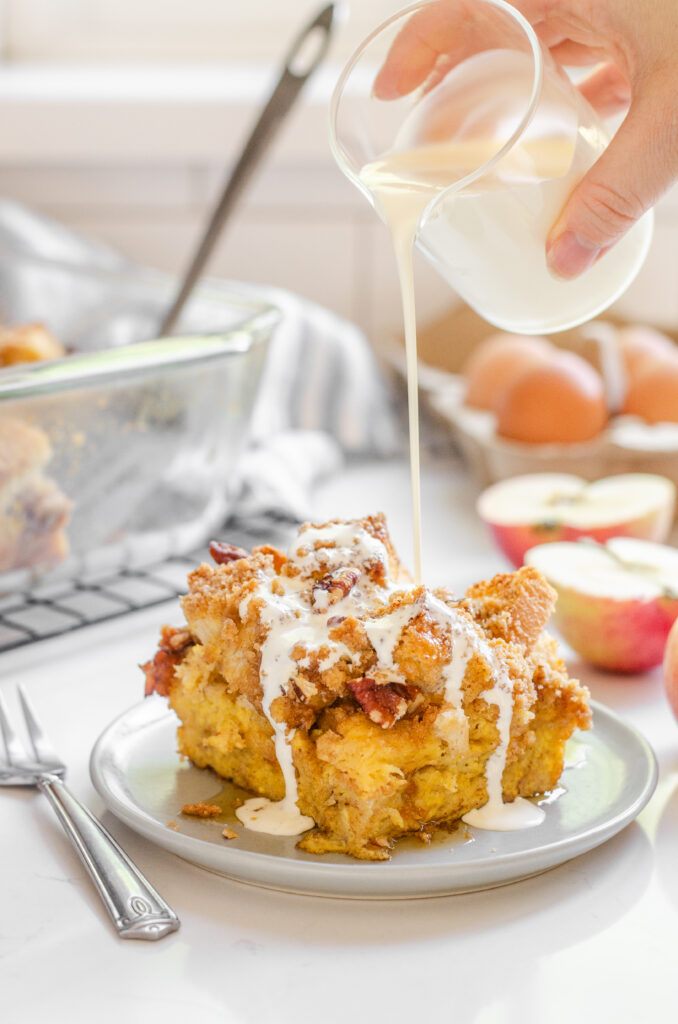 A slice of apple French toast casserole on a grey plate with apples in the background and cream pouring on top.