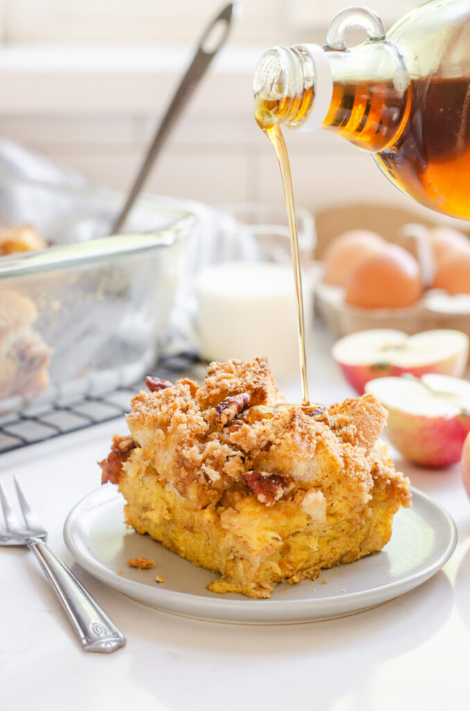 A slice of apple French toast casserole on a grey plate with apples in the background and maple syrup pouring on top.