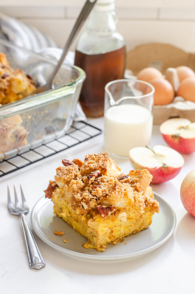 A slice of apple French toast casserole on a grey plate with apples in the background.