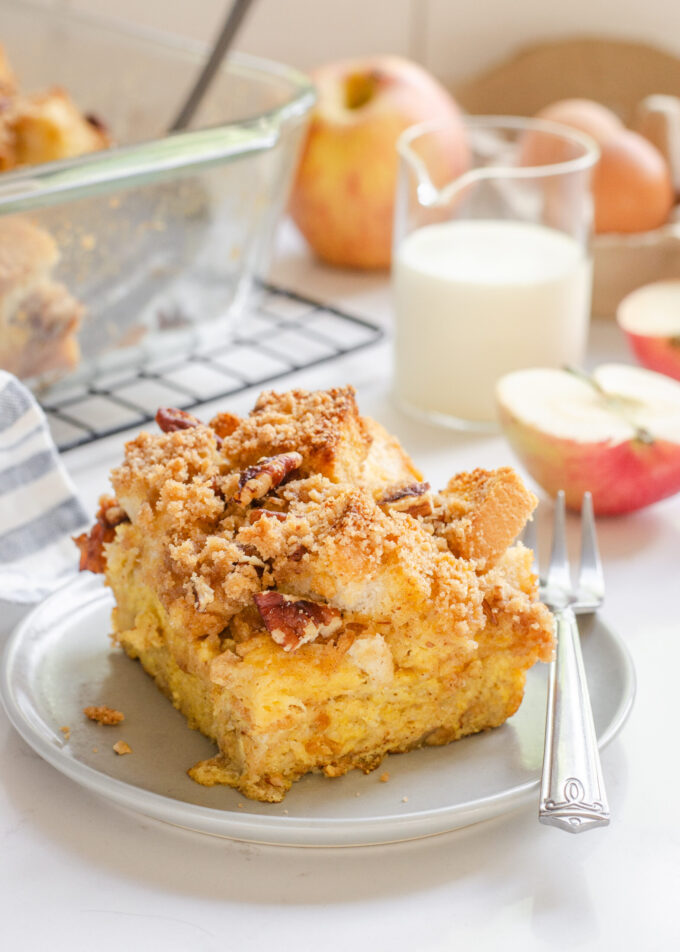 A slice of apple French toast casserole on a grey plate with apples in the background.